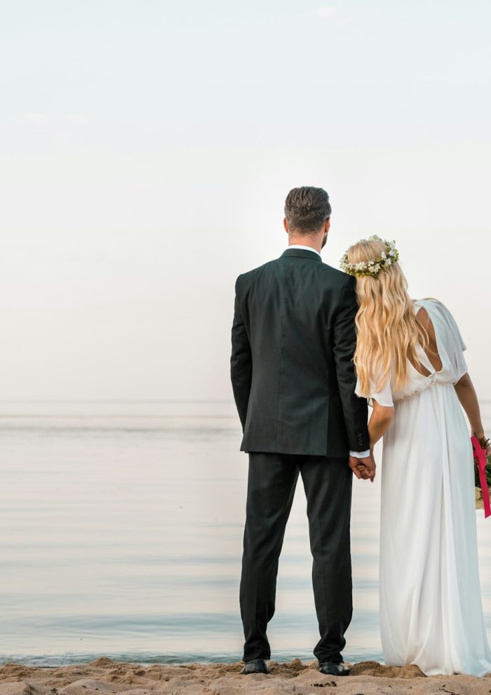 back view of wedding couple standing on beach with wedding bouquet and looking at sea