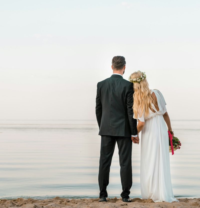 back view of wedding couple standing on beach with wedding bouquet and looking at sea